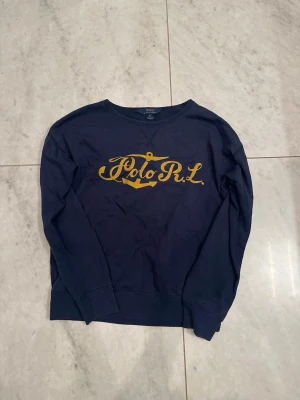 Marinblå stickad tröja Polo Ralph Lauren - Mörkblå stickad tröja från Polo Ralph Lauren med gul broderad logga och ankare på bröstet. Tröjan har rund halsringning och långa ärmar. Perfekt för dig som gillar klassisk och sportig stil. Passar S-M