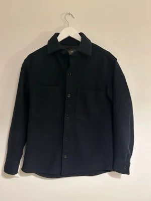 Mörkblå overshirt från H&M - Stilren mörkblå overshirt från H&M med klassisk krage och raka ärmar. Två stora bröstfickor framtill och knäppning med svarta knappar. Tillverkad i ett tjockare tyg som ger en schysst och avslappnad look. Perfekt för en lite kallare dag när man vill se lite schysstare ut. 