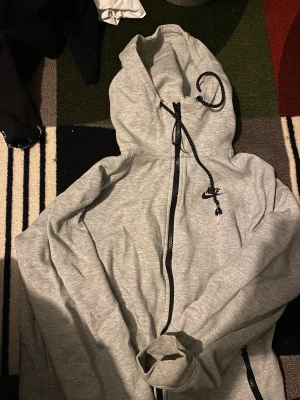 Grå zip hoodie från Nike - Snygg grå zip hoodie från Nike med svart logga på bröstet. Hoodien har dragkedja framtill, justerbar huva med svarta snören och fickor på sidorna. Perfekt för en avslappnad och sportig stil. Tillverkad i mjukt sweatshirtmaterial. Det är två olika modeller 