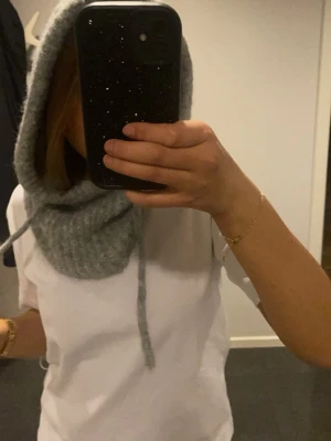 Grå stickad balaklava med snören - Säljer en mjuk och varm grå stickad balaklava med långa snören framtill. Perfekt för kalla dagar och ger en cool, avslappnad look. Passar dig som vill ha något trendigt och bekvämt till vintern. Den är beige nt grå men samma modell