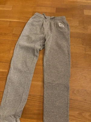 Gråa mjukisbyxor från Tracks - Säljer ett par klassiska gråa mjukisbyxor från Tracks med resår i midjan och dragsko. Byxorna har raka ben och sidofickor, samt en liten logga på vänster sida. Perfekta för chill dagar eller träning.