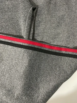 Grå mjukisbyxor med röd och svart rand - Snygga grå mjukisbyxor med broderad S.F-logga på benet. Byxorna har en röd och en svart rand längs sidan som ger en sportig vibe. Tillverkade i mjukt och stretchigt material för maximal komfort. Perfekta för chill eller träning.