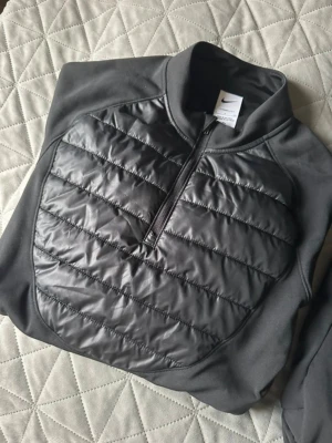 Nike träningsset svart halvzip & byxor - Säljer ett svart träningsset från Nike med halvzip-tröja och matchande byxor. Tröjan har vadderad framsida i glansigt material och dragkedja, medan byxorna har elastisk midja och orange Nike-logga. Perfekt för träning eller chill. Pris kan ej diskuteras 