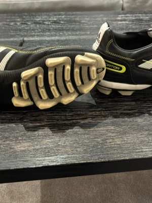 Adidas Predator svarta fotbollsskor - Säljer ett par klassiska Adidas Predator fotbollsskor i svart med vita och limegröna detaljer. Skorna har de ikoniska tre ränderna på sidan, snörning och robusta dobbar för bra grepp på planen. Ovandelen är i syntetmaterial och designen är sportig och stilren. Dom är storlek 42 och 2/3 passar även 43, priset på dessa nya ligger på runt 3000kr