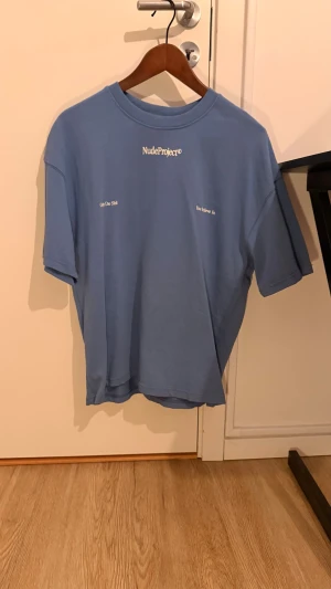Blå Nude Project t-shirt med tryck - Säljer en blå t-shirt från Nude Project med stort grafiskt tryck och text på ryggen samt mindre tryck framtill. T-shirten har normal passform, rund hals och är kortärmad. Perfekt för dig som gillar streetwear och statement-plagg.