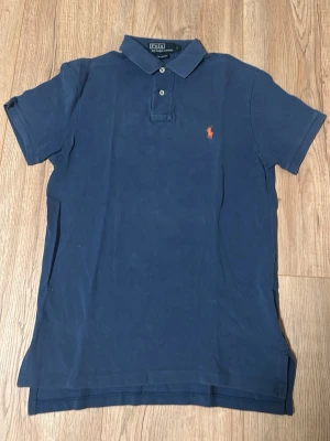 Marinblå pikétröja från Ralph Lauren - Klassisk marinblå pikétröja från Ralph Lauren med orange broderad logga på bröstet. Tröjan har korta ärmar, krage och två knappar framtill. Tillverkad i mjuk bomull och har en custom fit passform.