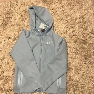 Ljusblå Nike nocta hoodie med dragkedja - Snygg ljusblå hoodie från Nike med dragkedja framtill och två fickor med dragkedja. Klassisk huva och diskret Nike-logga på bröstet. Perfekt för en sportig och avslappnad stil. Materialet känns mjukt och bekvämt.