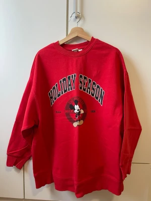 Röd Oversized Mickey Mouse Holiday Season tröja - Röd oversized sweatshirt med tryck 'Holiday Season' och en bild på Mickey Mouse framtill. Tröjan har rund hals, långa ärmar och ribbade muddar. Perfekt för dig som gillar Disney och vill ha en färgstark och bekväm look.