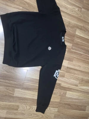 Svart sweatshirt med tryck på ärmen - En svart sweatshirt med rund hals och tryck på vänster ärm samt ett litet märke på bröstet. Tröjan har en avslappnad passform och ribbade muddar vid ärmslut och nederkant. Perfekt för en casual look. Står XL men sitter som M/L 