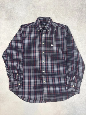 Rutig långärmad skjorta Burberry - Klassisk rutig skjorta från Burberry i mörkblå, röd och vit. Skjortan har lång ärm, knappar framtill och en liten broderad logga på bröstet. Tillverkad i mjuk bomull och har en normal passform. Perfekt för en stilren och avslappnad look.