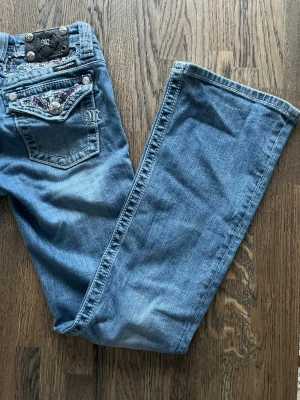 Miss Me bootcut jeans med detaljer - Snygga blå bootcut jeans från Miss Me med dekorativa bakfickor som har pärl- och paljettbroderier samt silverfärgade knappar. Klassisk femficksmodell i mjuk denim med coola detaljer och logga på bakfickan. Perfekta för dig som gillar statement-jeans.