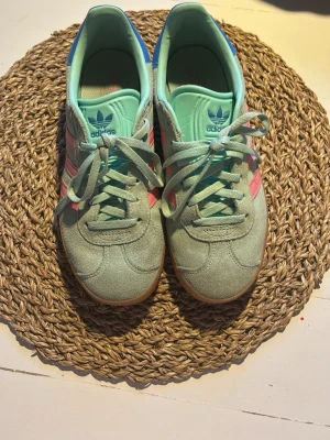 Adidas Gazelle mintgrön/rosa sneakers - Säljer ett par Adidas Gazelle sneakers i mintgrönt mocka med rosa ränder och blå detalj på hälen. Skorna har klassisk gummisula och lågt skaft. Perfekta för dig som vill sticka ut med färg och retrostil.