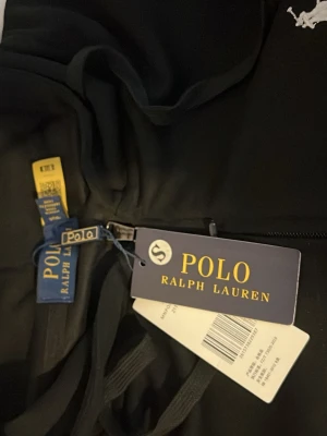 Svart hoodie från Polo Ralph Lauren - Svart hoodie från Polo Ralph Lauren med dragkedja framtill och klassisk huva med dragsko. Ikoniska vita Polo-loggan broderad på bröstet. Tillverkad i mjukt material som känns skönt mot huden. Perfekt för en avslappnad och stilren look.
