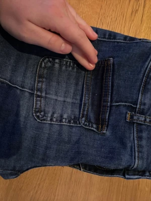 Blå jeans med fickdetalj - Säljer ett par blå jeans med unik fickdetalj på sidan och kontrastsömmar. Jeansen har raka ben och klassisk midja. Perfekta för dig som vill ha något lite annorlunda i garderoben.
