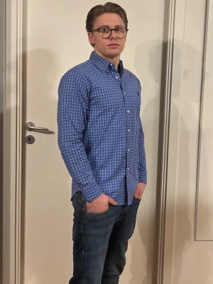 Blå rutig Ralph Lauren skjorta  - Snygg blå skjorta med vitt rutmönster och klassisk krage. Skjortan har långa ärmar och knäppning framtill. Perfekt för en stilren och avslappnad look.