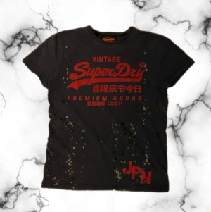 Svart Superdry t-shirt med röd print - Fet svart t-shirt från Superdry med röd vintage-logga och japanska tecken på bröstet. T-shirten har korta ärmar och är dekorerad med unika färgstänk över hela plagget. Passar dig som har lite smalare axelbredd. 