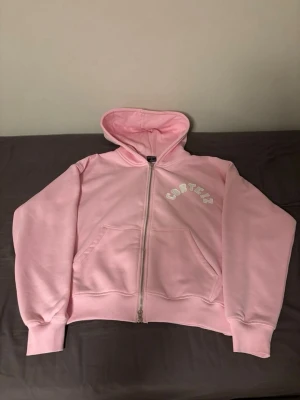  hoodie  Corteiz - Säljer en ljusrosa hoodie från Corteiz med dragkedja och stor huva. Framtill står Corteiz broderat i vitt på bröstet och stort på ryggen. Hoodien har kängurufickor och ribbade muddar. Perfekt för en chill och trendig look.