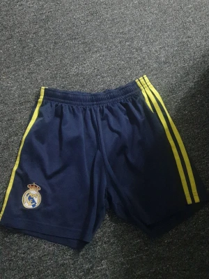 Real Madrid blå träningsshorts - Mörkblå träningsshorts från Adidas med Real Madrid-logga på vänster ben och gula ränder längs sidorna. Shortsen har elastisk midja och är tillverkade i ett lätt och bekvämt syntetmaterial, perfekta för fotboll eller träning.