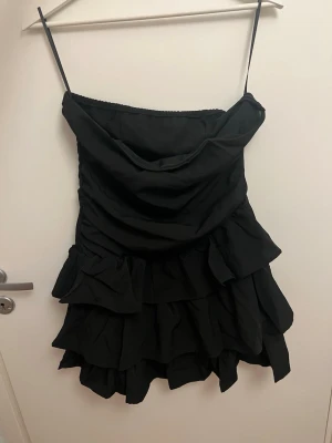 Svart volangklänning med tunna band - Supersnygg svart klänning med tunna axelband och flera lager volanger. Draperad överdel som ger en cool look och kort, figurnära passform. Perfekt för dig som gillar trendiga detaljer och vill sticka ut. Materialet känns lätt och följsamt.
