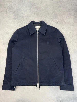 Svart jacka från Ami Paris med dragkedja - Stilren svart jacka från Ami Paris med klassisk krage och silverfärgad dragkedja framtill. Jackan har två snedställda fickor, diskret Ami-logga på bröstet och justerbara detaljer baktill. Perfekt för dig som gillar minimalistisk design.