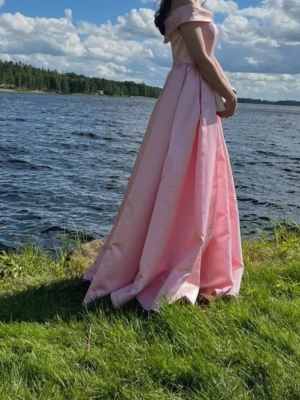  balklänning - Elegant ljusrosa balklänning med offshoulder-design och vid, golvlång kjol. Klänningen har en klassisk, figurnära överdel och är tillverkad i ett glansigt material som ger en lyxig känsla. Perfekt för bal eller student.