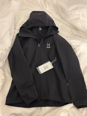 Svart vindjacka från Haglöfs - Helt ny köpt på xxl för 1750kr Snygg svart vindjacka från Haglöfs med dragkedja framtill och justerbar huva. Jackan har minimalistisk design med logga på bröstet och är tillverkad i ett lätt, vindtätt material. 