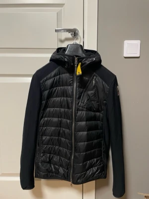 Svart hybridjacka med huva Parajumpers - Snygg svart hybridjacka från Parajumpers med quiltad front och rygg i glansigt material samt stickade ärmar och sidor. Jackan har huva, bröstficka med dragkedja och gul detalj vid halsen. Perfekt för lager-på-lager och streetwear. Pris kan diskuteras 