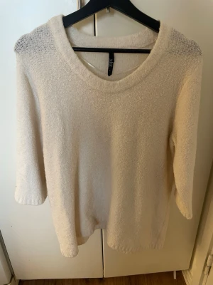 Krämvit stickad tröja från Vila - Mysig krämvit stickad tröja med rund halsringning och trekvartsärmar. Tröjan har en lätt luddig struktur och är tillverkad i ett mjukt material som känns skönt mot huden. Perfekt för lager på lager och enkel att matcha.