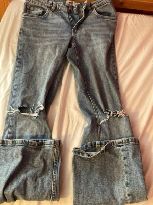 Bootcut jeans från 157 med slitningar - Säljer ett par ljusblå bootcut jeans från 157 med slitna detaljer vid knäna och breda uppvikta benslut. Klassisk femficksmodell i denim med låg midja. Perfekta för dig som gillar en avslappnad och trendig stil. Dem blev försmå när jag tvättade dem 🧼 