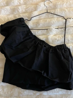 Svart oneshoulder topp med volang - Trendig svart oneshoulder topp med stor volang längs halsringningen. Perfekt för dig som vill sticka ut med en cool och modern look. Toppen är ärmlös och har en snygg asymmetrisk design.💓‼️mindre än vad storleken säger passar mig bra som normalt bär s/m‼️