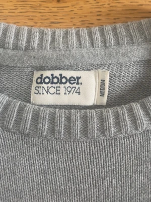 Ljusgrå stickad tröja från Dobber - Snygg ljusgrå stickad tröja från Dobber med rund halsringning och ribbade muddar vid ärmslut och nederkant. Enkel och stilren design som passar perfekt till jeans eller chinos. Skön och mjuk känsla, perfekt för kyligare dagar.