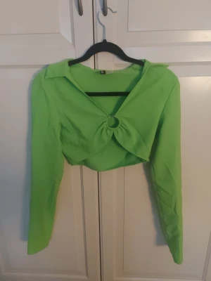 Limegrön croppad topp med ringdetalj - Trendig limegrön croppad topp med lång ärm och snygg ringdetalj framtill. Toppen har en djup v-ringning och är tillverkad i ett mjukt syntetmaterial. Perfekt för dig som vill sticka ut med en färgstark look.