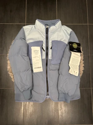 Ljusblå dunjacka från Stone Island - Snygg ljusblå dunjacka från Stone Island med hög krage och dragkedja framtill. Jackan har paneler i olika blå nyanser och detaljer i mesh-tyg upptill. Klassisk Stone Island patch på ärmen och två stora fickor framtill. Perfekt för kalla vinterdagar. Kvittot finns och medkommer vid köp! 