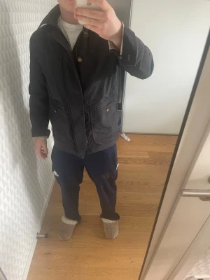 Svart rock med krage och knappar - Klassisk svart rock med bred krage och knäppning framtill. Jackan har stora fickor med lock och knappdetaljer. Passformen är normal och materialet ser ut att vara bomull eller polyester, perfekt för kyligare dagar.