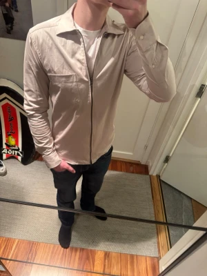 Beige overshirt från John Henric - Stilren beige overshirt från John Henric med dragkedja framtill och en bröstficka. Skjortkrage och långa ärmar med knapp vid ärmslut. Tillverkad i ett tunt, mjukt bomullstyg som passar perfekt för lager på lager.
