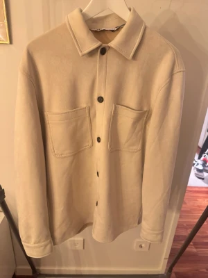 Beige overshirt med stora fickor - Snygg beige overshirt med raka linjer och två stora bröstfickor. Stängs med svarta knappar framtill och har klassisk krage. Materialet ser ut att vara mjukt och lite tjockare, perfekt för lager på lager. Passar till både hoodie och t-shirt.