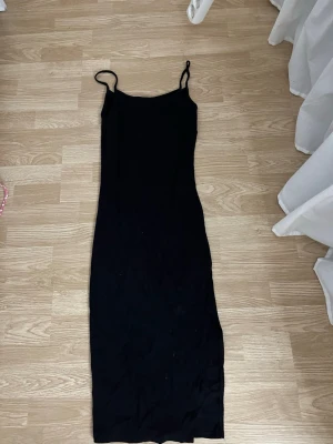 Svart långklänning med smala axelband - En stilren svart långklänning med smala axelband och figurnära passform. Klänningen har en enkel design utan mönster och är perfekt för dig som gillar minimalistisk stil. Materialet känns mjukt och följsamt mot kroppen.