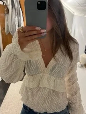 Vit omlottopp med volang från Ginatricot  - Säljer en vit omlottopp från Ginatricot med lång ärm och v-ringning. Toppen har ett vågigt strukturmönster och volangdetalj framtill. Perfekt att styla med jeans eller kjol för en trendig look💕💕