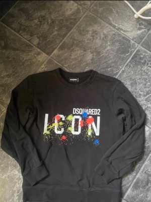 Svart ICON sweatshirt från Dsquared2 - Säljer en svart sweatshirt från Dsquared2 med stor vit 'ICON'-text framtill och färgglada färgstänk i rött, blått och gult. Tröjan har rund halsringning och ribbade muddar vid ärmar och nederkant. Perfekt för dig som gillar streetwear och statementplagg.