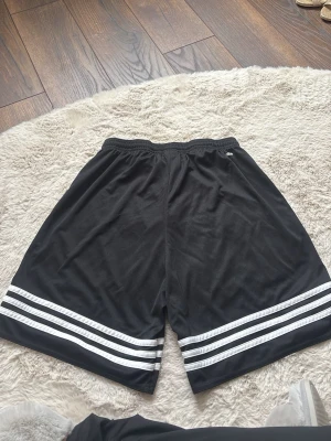 Svarta Adidas shorts med vita ränder - Säljer ett par svarta Adidas shorts med klassiska vita ränder längs benen. Shortsen har elastisk midja med snörning och är tillverkade i ett lätt och svalt syntetmaterial, perfekta för träning eller sportiga dagar.