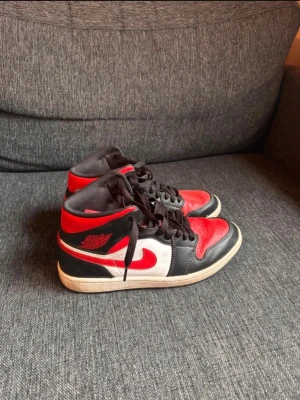 Air Jordan 1 Retro svart/röd/vit - Säljer ett par klassiska Air Jordan 1 Retro sneakers i svart, rött och vitt läder. Skorna har högt skaft, svart snörning och ikoniska röda detaljer med vinge-loggan på sidan. Perfekta för dig som gillar streetwear och vill sticka ut med en tidlös modell.