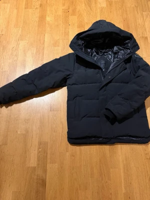 Canada Goose Macmillan (pris kan diskuteras) - Snygg svart dunjacka med stor huva och quiltad design. Jackan har en patch på ärmen och dragkedja med knappslå framtill. Perfekt för kalla vinterdagar och har en modern, boxig passform. Pris kan diskuteras vid seriös intresse. Den är true to size.