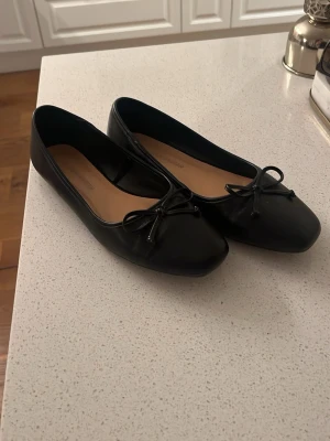 Svarta ballerinaskor med rosett - Stilrena svarta ballerinaskor med klassisk rund tå och söt rosettdetalj framtill. Skorna har en låg profil och är tillverkade i ett slätt, glansigt material med beige innersula. Perfekta för en enkel och trendig look.
