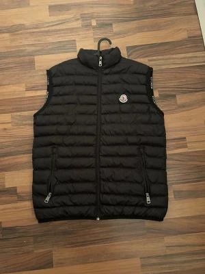 Svart dunväst från Moncler - Snygg svart dunväst från Moncler med quiltad design och hög krage. Västen har två dragkedjefickor framtill och Moncler-logga på bröstet. Perfekt för lager-på-lager och håller dig varm med stil. Klassisk passform och diskreta detaljer.