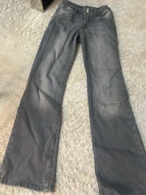 Grå bootcut jeans med låg midja - Säljer ett par grå jeans med bootcut passform och låg midja. Jeansen har klassiska fem fickor och dragkedja med knapp framtill. Perfekta för en avslappnad och trendig look.