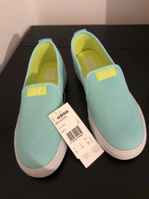 Turkosa slip-on sneakers från Adidas - Fräscha slip-on sneakers från Adidas NEO i turkos canvas med vita sulor och ljusgula detaljer. Skorna har elastiska paneler för enkel påtagning och en sportig vibe med gula ränder bak. Perfekta för en chill och trendig stil.