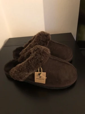 Bruna fårskinnstofflor från Shepherd - Mysiga bruna tofflor från Shepherd i äkta fårskinn med fluffig insida och kant. Perfekta att glida runt i hemma när det är kallt. Klassisk slip-in modell med mjuk sula och naturligt material som andas.