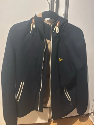 Marinblå vindjacka från Lyle & Scott - Snygg marinblå vindjacka från Lyle & Scott med dragkedja, vita detaljer på fickor och snören samt gul logga på bröstet. Jackan har huva med dragsko och är perfekt för blåsiga dagar. Klassisk och stilren design som funkar till det mesta.