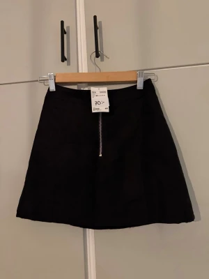 Svart kortkjol med dragkedja H&M - Svart kortkjol från H&M med rak passform och en snygg silverfärgad dragkedja framtill. Kjolen är enkel och stilren, perfekt att matcha med olika toppar. Materialet ser ut att vara mocka eller syntet med mockakänsla. Ca 40cm