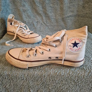 Vita Converse All Star high tops - Klassiska vita Converse All Star high tops med canvasöverdel, gummisula och ikonisk logga på sidan. Skorna har vita snören, metallöljetter och svart rand runt sulan. Perfekta för en avslappnad och tidlös streetstil.